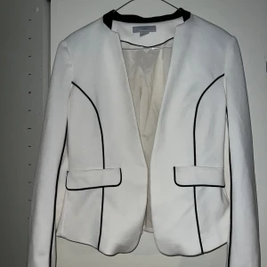 Supersnygg kavaj - Säljer min jättefina kavaj, som jag tyvärr inte kan ha längre. Den har två fläckar (se bild). Figursydd. Vit med svarta detaljer. Guldkedja.