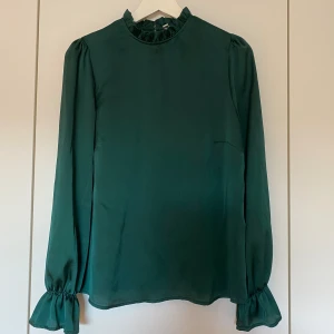 Mörkgrön blus - Mörkgrön blus i glansigare material. Lite volanger vid kragen samt nedtill vid armarna. Storlek 34 från Nelly, passar även som 36 då den är lite större i storleken 