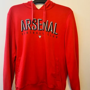Arsenal tröja - Arsenal tröja i röd färg med blå logga. I storlek medium. Passar både tjej och kille. Fint material med skön passform. Använd ett fåtal gånger men har en liten tråd som sticker ut på tröjan som en defekt. Ny köp priset var 950kr.