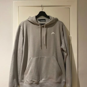 J.Lindeberg - En grå J.Lindeberg hoodie med en liten vit logga på bröstet. Huvan är fodrad med mjukt tyg och har en dragsko för att justera passformen. Hoodie har en ficka på magen.