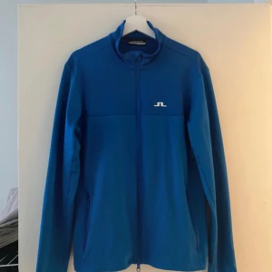 J.Lindeberg - En blå jacka med J.Lindeberg-logotypen på bröstet och baksidan. Jackan har en dragkedja framtill och två fickor på sidorna.