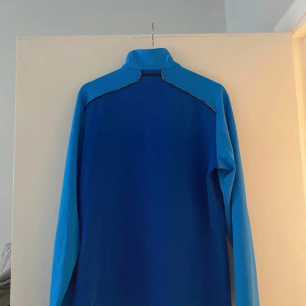 En blå tröja med en halv dragkedja i halsen. Tröjan har en mörkblå färggradient på vänster sida och en ljusare blå färggradient på höger sida. Tröjan har en liten J.Lindeberg-logotyp på vänster bröst.. Sport & träning.
