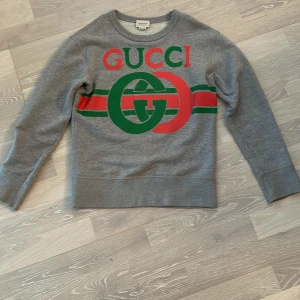 En riktigt snygg Gucci tröja perfekt för våren,  - 39 cm i midjan.   46,5 cm i ärmen.  55 cm längd Strlek 10 år i barn Färg: grå  Material: 100% bomull