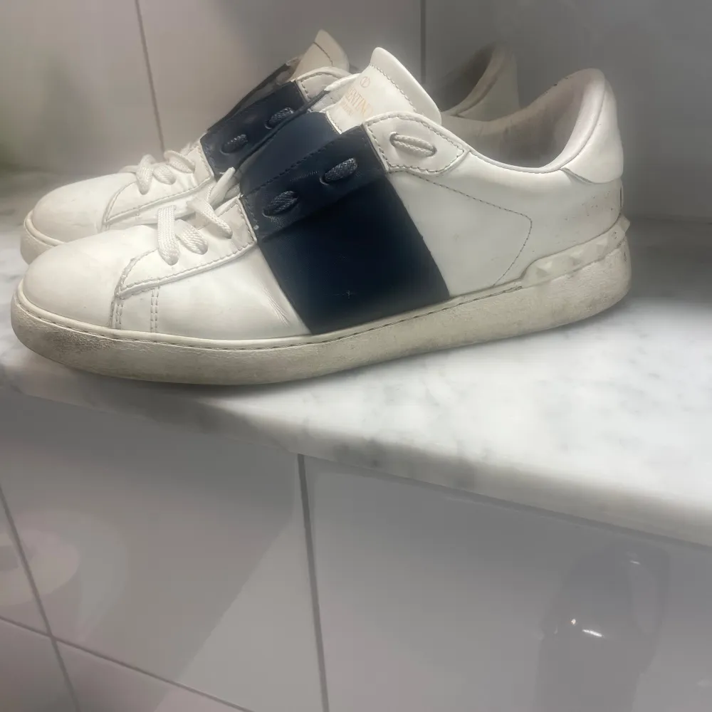 Ett par vita sneakers från Valentino med en bred mörkblå rem över snörningen. Skorna har en rund tå och en vit sula.storlek 39 passar 0,5 större . dustsbag tillkommer. Skor.