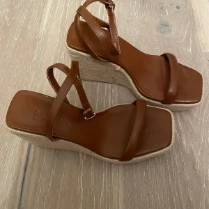 Bruna sandaler med kilklack - Ett par bruna sandaler med kilklack och remmar. Sandalerna har en fyrkantig tå och en justerbar rem runt ankeln.