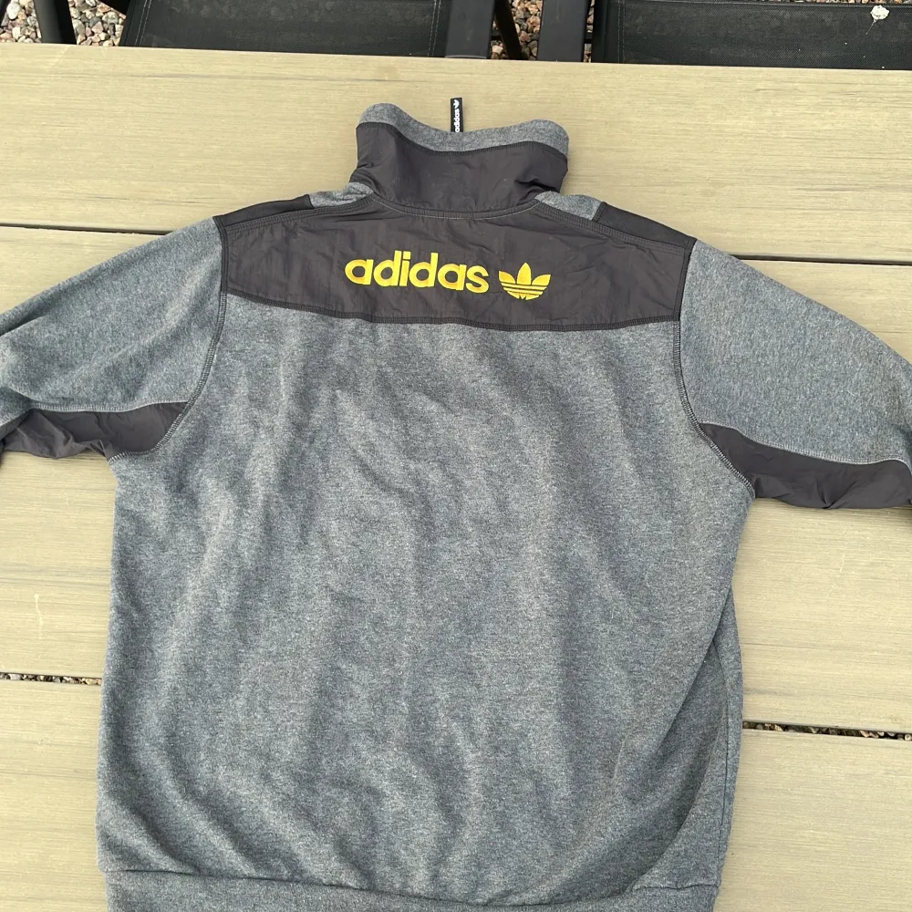 En grå och svart tröja från Adidas med gul logotyp på bröstet och ryggen. Tröjan har en halv dragkedja framtill och är tillverkad i ett mjukt material. Den har långa ärmar och ribbstickade muddar vid ärmslut och nederkant.. Tröjor & Koftor.