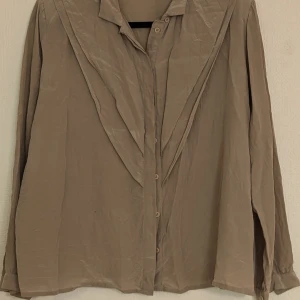 Beige blus i 100% siden - En elegant beige blus från Crêpe de Chine tillverkad i 100% siden. Blusen har långa ärmar och knappar framtill. Den har en lös passform och en unik krage. lite mindre i storlek
