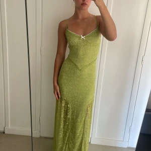 Grön långklänning från Tiger Mist - En elegant grön långklänning från Tiger Mist med smala axelband och en liten vit rosett framtill. Klänningen har ett subtilt mönster och är tillverkad i ett lätt och bekvämt material.