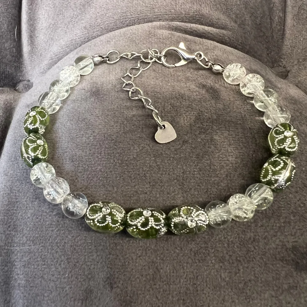 Ett elegant armband med gröna och genomskinliga pärlor. De gröna pärlorna har silvriga dekorativa mönster. Armbandet har en justerbar kedja mellan 17-22 cm och avslutas med med ett hjärtformat hänge.. Asusteet.