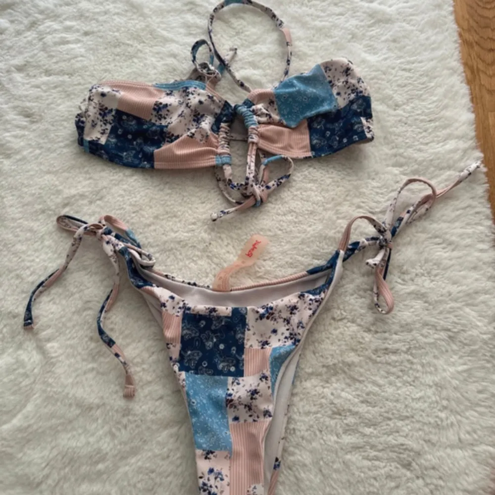 En färgglad bikini med blommigt mönster i blått, rosa och vitt. Överdelen har knytband och en snörning framtill. Underdelen har knytband på sidorna.. Övrigt.