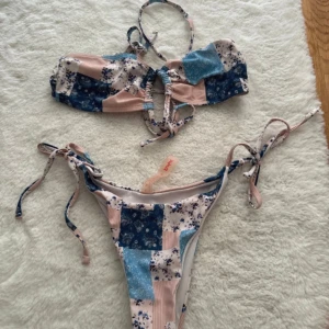 Blommig bikini med knytband - En färgglad bikini med blommigt mönster i blått, rosa och vitt. Överdelen har knytband och en snörning framtill. Underdelen har knytband på sidorna.