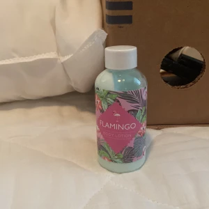 Flamingo Body Lotion - En kroppslotion från Flamingo med en färgglad etikett som har ett tropiskt mönster med gröna blad och rosa detaljer. Flaskan är genomskinlig med en vit kork.