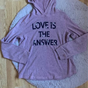 Rosa hoodie med texten 'LOVE IS THE ANSWER' - En bekväm rosa hoodie med texten 'LOVE IS THE ANSWER' på framsidan. Tröjan har långa ärmar och ribbstickade muddar vid ärmslut och nederkant. Den är tillverkad i ett mjukt material som passar perfekt för kyligare dagar.
