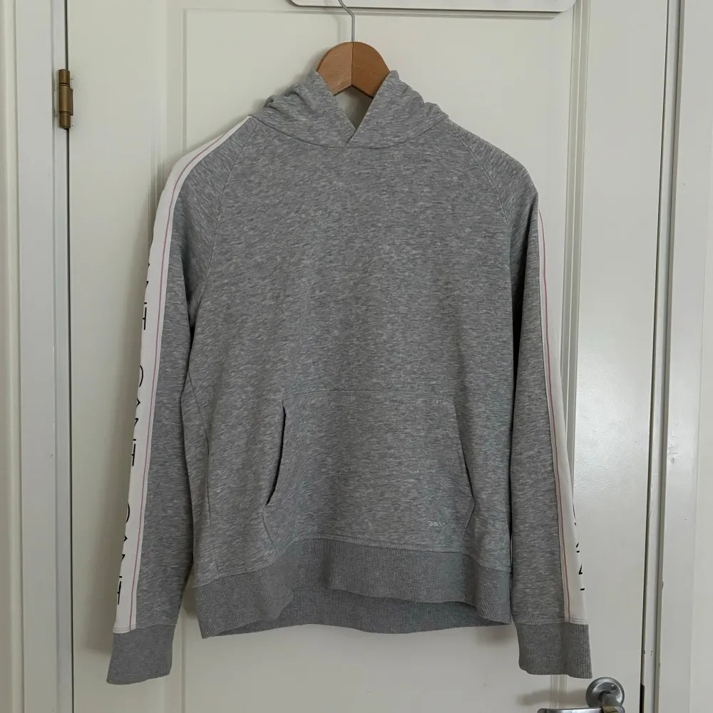Detta är en grå hoodie av märket Gant. Den har svarta, rosa och vita inslag längst båda ärmarna. Hoodien är i gott skick utan några defekter!. Hupparit & Collegepaidat.