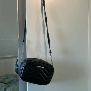 Axelväska - En svart väska i fake skinn 20x13cm stor, långt axelband med liten silver kedja. Använt fåtal gånger, är i bra skick.