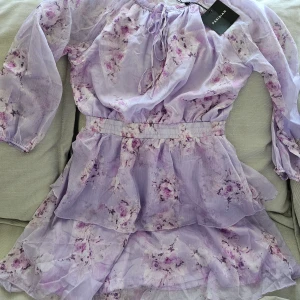 Lila blommig klänning från Parisian  - En lila blommig klänning från Parisian med långa ärmar och volangdetaljer. Klänningen har en elastisk midja och ett knytband vid halsen. Materialet är lätt och genomskinligt med ett innerfoder för extra komfort.