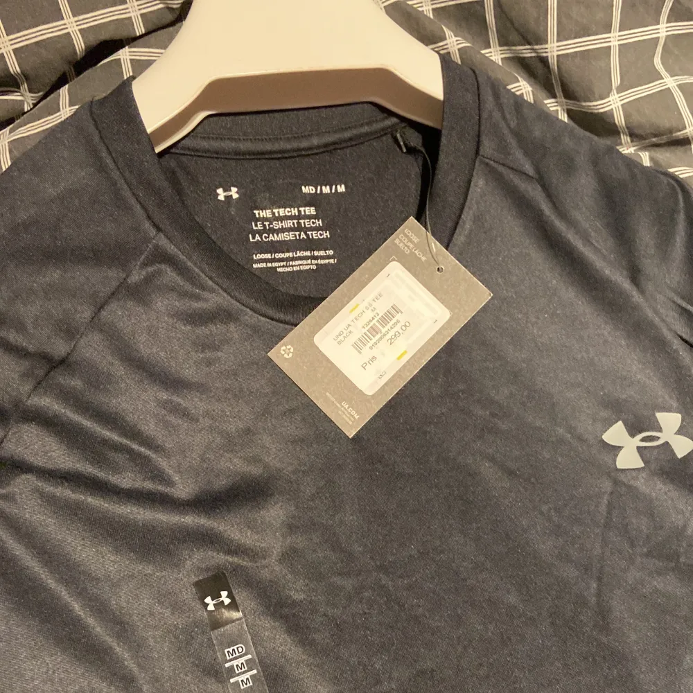 En svart t-shirt från Under Armour med korta ärmar och rund hals. T-shirten har en liten logotyp på bröstet och en större logotyp på ryggen. Den är tillverkad i ett lätt och andningsbart material, perfekt för träning.. T-shirts.