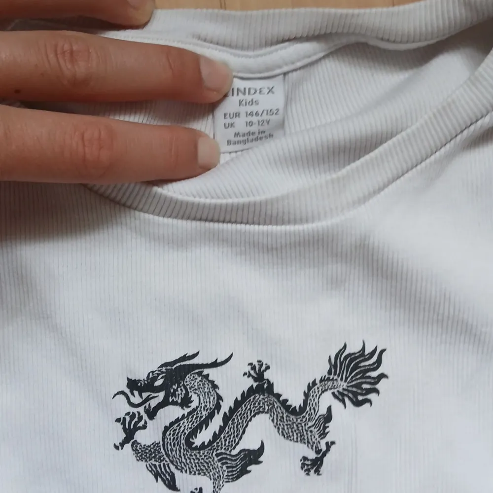 En vit långärmad tröja från Lindex med ett svart drakmönster på bröstet. Tröjan har ribbade muddar vid ärmslut och nederkant. Obs barnstorlek men sitter som en XS om man gillar baby tee looken.. Neuletakit & Villapaidat.
