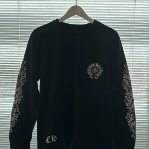 Chrome Hearts - Denna långärmade tröja från Chrome Hearts är svart med vita detaljer och märkets logotyp på bröstfickan. Tröjan har dekorativa mönster längs ärmarna och är tillverkad av högkvalitativt material. Ett exklusivt plagg i utmärkt skick! Priset kan diskute