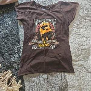 Brun t-shirt från Skank - En brun t-shirt från Skank med ett tryck på framsidan som visar en knuten näve och texten 'Show Your Lion Paw'. T-shirten har korta ärmar och en rund halsringning.