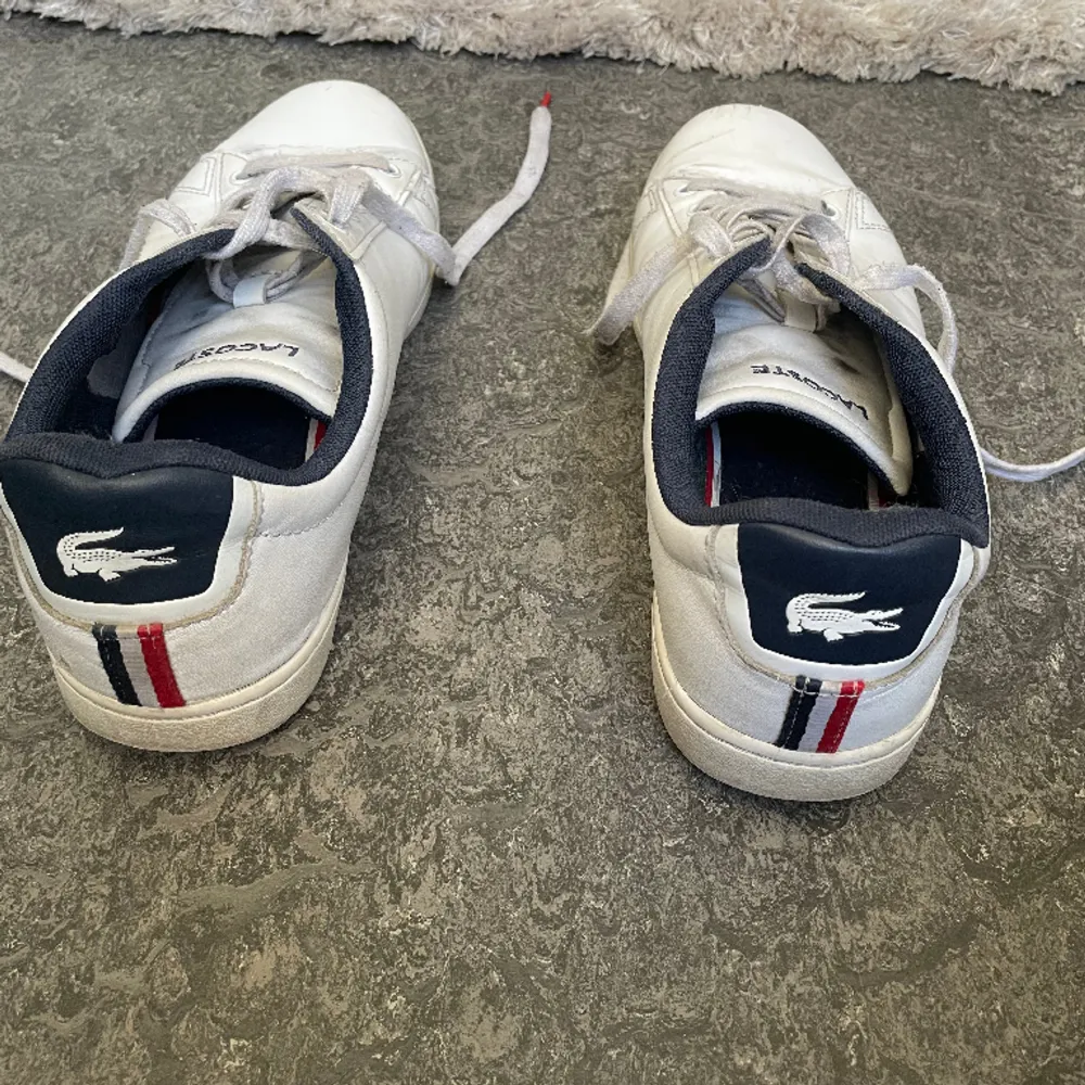 Här har du chansen att köpa ett par använda Lacoste Sneakers Carnaby i storlek 42 till ett bra pris! Skorna kommer i en stilren design med vitt som bas och fina detaljer i marinblått och rött. Trots några små skador är skorna fortfarande i gott skick. Skor.