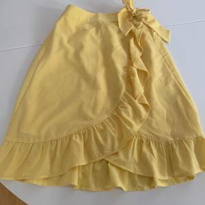Gul medellång kjol med en rosett på ena sidan i midjan och volnang i nedre kanten och snett över från rosetten till nedre kanten från Lindex. Använd fåtal gånger så i fint skick. Storlek 134/140. 