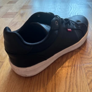 Svarta sneakers från Levi's - Ett par svarta sneakers från Levi's med vita sulor och snörning. Skorna har en röd Levi's-logga på sidan och är tillverkade i skinn.