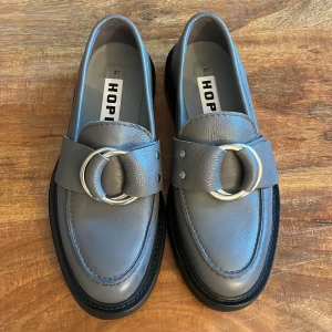 Grå loafers från HOPE - Ett par grå loafers från Hope i storlek 37. Skorna har en dekorativ rem med en stor silverfärgad spänne på ovansidan. De är tillverkade i skinn och har en svart sula. Aldrig använda då det sitter för tight. Ordinariepris: 3500kr 