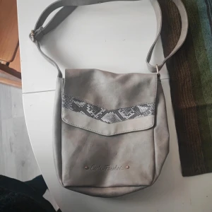 Grå axelväska från Carla Faustini - En stilren grå axelväska från Carla Faustini med ett dekorativt mönster i ormskinnsdesign på framsidan. Väskan har en justerbar axelrem och en dragkedja för säker förslutning. Insidan har flera fack för organisering.