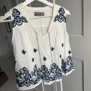 Vit söt topp - Vitt linne med blåa mönsterdetaljer, samt snören att knyta framtill. Storlek XS. OBS skrynklig på bilden.