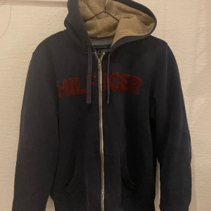 Marinblå zip hoodie Tommy Hilfiger - Marinblå huvtröja från Tommy Hilfiger med dragkedja, fickor och beige fluff inuti. Storlek M. Använd men i gott skick. Nött på muddarna, se bild tre. Inköpspris 850kr