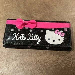 Hello Kitty plånbok - Glittrig plånbok med Hello Kitty-motiv. Söt rosa rosett. Fack för kort, mynt samt kvitton och pengar. Mått 18,5x9 cm. 