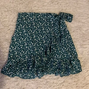 Volangkjol - Somrig kjol som är grön med vita blommor och har volanger. Har knappar i midjan (på sidan) och även ett band som man kan knyta till en söt rosett. Använd en eller två gånger, därav bra skick. Säljer pga att den inte passar. 40kr plus frakt!!💕💕💕