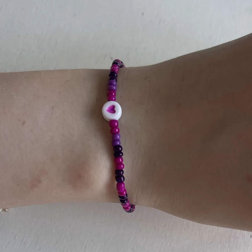 Ett egengjort pärlarmband med färgerna magenta, mörklila och en ännu mörkare lila. Armbandet har en annan pärla i vit som har en rosa hjärtsymbol på. . Accessoarer.