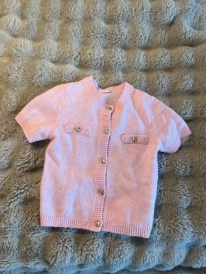 Ljusrosa kortärmad kofta - Söt ljusrosa kofta med korta ärmar och guldfärgade knappar framtill. Två dekorativa fickor med knappdetaljer på bröstet. Ribbstickade kanter vid ärmslut, hals och nederkant. Perfekt för en fin och söt look.💖