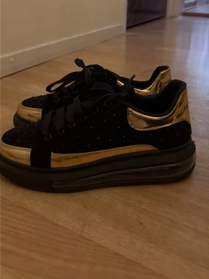 Svarta och guldiga sneakers Fashion - Helt oanvända snygga sneakers i svart och guld med glittrande detaljer och bred svart sula. Skorna har svarta skosnören och metallic guldpaneler på sidan och hälen. Ovandelen är dekorerad med små svarta stenar.  Pris kan absolut diskuteras . 
