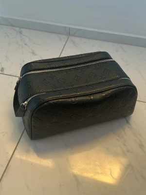 Svart necessär från Louis Vuitton - Snygg svart necessär från Louis Vuitton i präglat läder med klassiskt monogrammönster. Väskan har två stora fack med silverfärgade dragkedjor och en praktisk bärrem på sidan. Perfekt för att organisera dina essentials på resan.
