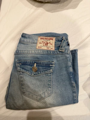 True Religion Billie ljusblå jeans - Ljusblå jeans från True Religion, modell Billie Mid Rise Straight. Klassisk femficksdesign med snygga detaljer och emblem på bakfickan. Jeansen är raka i modellen och har en mellanhög midja. Materialet är denim och passar perfekt till en avslappnad look.