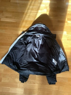 PLEIN SPORT pufferjacka med vita ränder - Snygg svart pufferjacka med huva och vita ränder längs ärmarna. Jackan har dragkedja framtill och två sidofickor. Perfekt för kalla dagar och ger en sportig look. Materialet är glansigt och vadderat för extra värme.