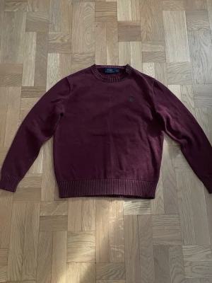 Vinröd stickad tröja Polo Ralph Lauren - Vinröd stickad tröja från Polo Ralph Lauren med rund halsringning och ribbade muddar. Tröjan har en liten grön broderad logga på bröstet och är tillverkad i mjukt material. Perfekt för dig som gillar klassisk och stilren design. 