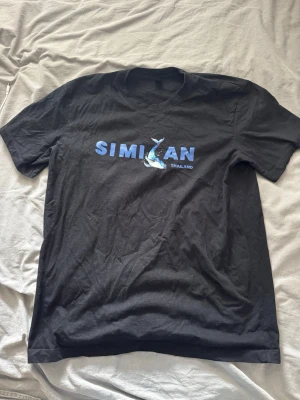 Svart Similan Thailand t-shirt - Cool svart t-shirt med blått Similan Thailand-tryck och en haj på bröstet. Klassisk rund hals och korta ärmar, perfekt för dig som gillar havet och äventyr. Materialet känns som bomull och passformen är normal.