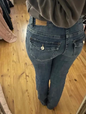 Blå bootcut jeans med fickdetaljer - Säljer ett par blå bootcut jeans med snygga fickor bak som har lock och silverknappar. Perfekta till sneakers eller boots för en avslappnad look.