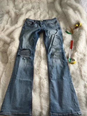 Blå bootcut jeans från ONLY - Säljer ett par ljusblå bootcut jeans från ONLY med slitningar och hål på ena benet. Jeansen har klassisk femficksmodell, låg midja och utsvängda ben. Perfekta för en avslappnad och trendig look. Midjemåttet är cirka 38cm och innerbenslängden 80cm