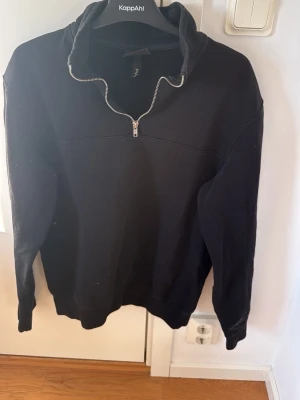 Svart halfzip hoodie från H&M - Svart hoodie från H&M med halv dragkedja framtill och hög krage. Tillverkad i mjukt sweatshirtmaterial som känns skönt mot huden. Klassisk och enkel design utan tryck eller mönster, perfekt till jeans eller joggers.