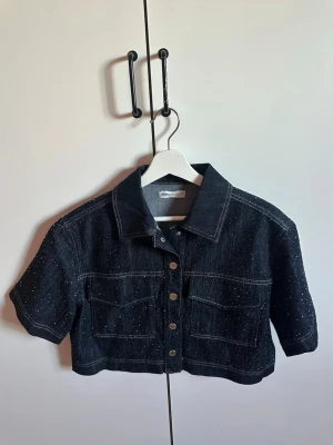 Kort jeansjacka med glitterdetaljer - Snygg mörkblå kort jeansjacka från gina tricot med korta ärmar, silvriga knappar och glitterprickar över hela tyget. Två stora bröstfickor framtill och markerade sömmar ger en cool och trendig look.