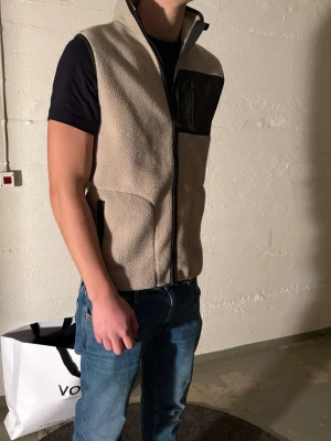 Beige fleeceväst med bröstficka - Snygg beige fleeceväst med hög krage och dragkedja framtill. Västen har svarta detaljer, inklusive en bröstficka med dragkedja och svarta kantband vid fickorna. Perfekt att slänga över en t-shirt för en chill look.
