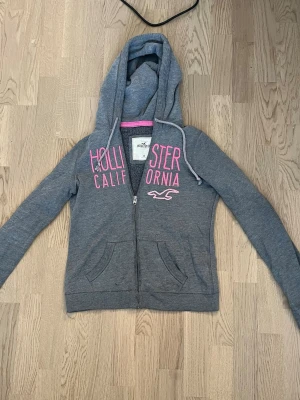 Grå zip hoodie från Hollister - Snygg grå hoodie från Hollister med dragkedja och huva. Stor rosa text 'Hollister California' och logga framtill. Mjuk bomullsblandning, två fickor fram och justerbara snören i huvan. Perfekt för en skön och cool outfit.