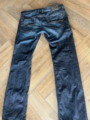 Mörkblå jeans från Diesel - Säljer ett par mörkblå jeans från Diesel med klassisk femficksmodell och snygga kontrastsömmar i orange. Jeansen har en rak passform och detaljerad bakficka med Diesel-logga på läderpatch bak i midjan. Perfekta för en avslappnad och trendig look.
