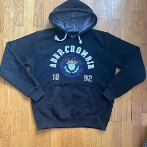 Svart Abercrombie & Fitch Vintage Hoodie - En supersnygg svart hoodie från Abercrombie and Fitch vintage-stil🤩Tröjan har ett tryck från Abercrombie med både text i tyg och dess logotyp👌En snygg och tjock tröja som denna passar perfekt nu under vintern❄️Tröjan är dessutom i mycket gott skick utan några defekter👍