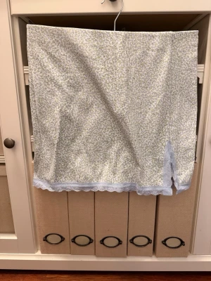 Blommig kjol med spets och slits - Söt kortkjol med småblommigt mönster i grönt och vitt. Kjolen har ljusblå spetsdetaljer längs nederkanten och en liten slits framtill som ger extra stil. Perfekt för dig som gillar romantiska och feminina plagg.